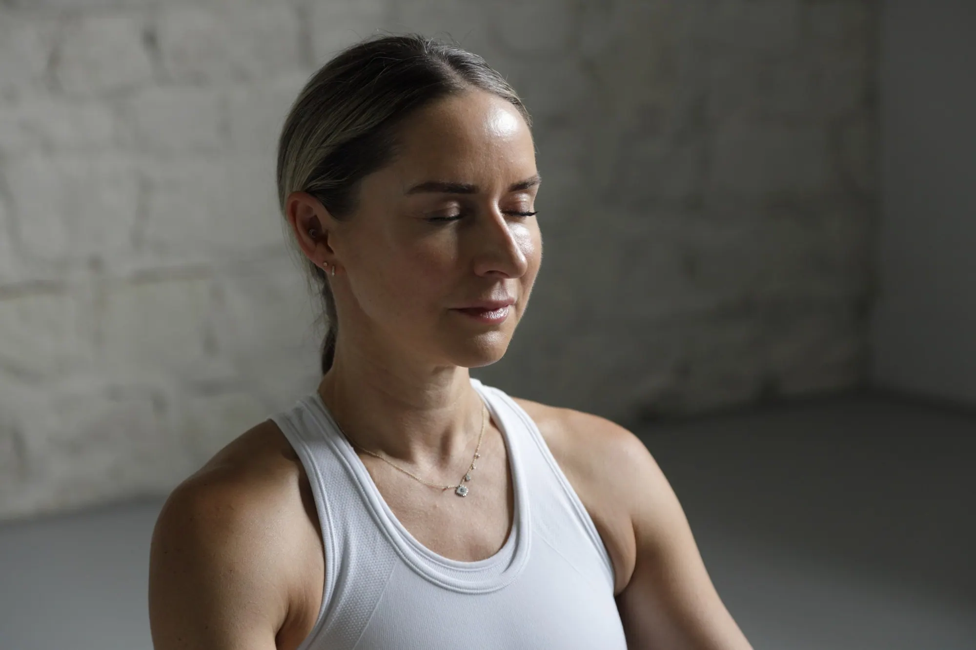 Chrissy in a white vest with her eyes closed, taking a moment before she moves, against the whitewashed brick of her studio.