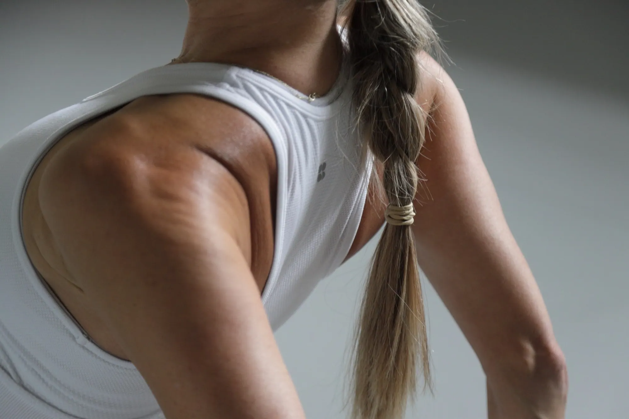 A close crop of Chrissy’s shoulder and upper back in a white vest, her long plait falling across it, in the cool grey light of her studio.