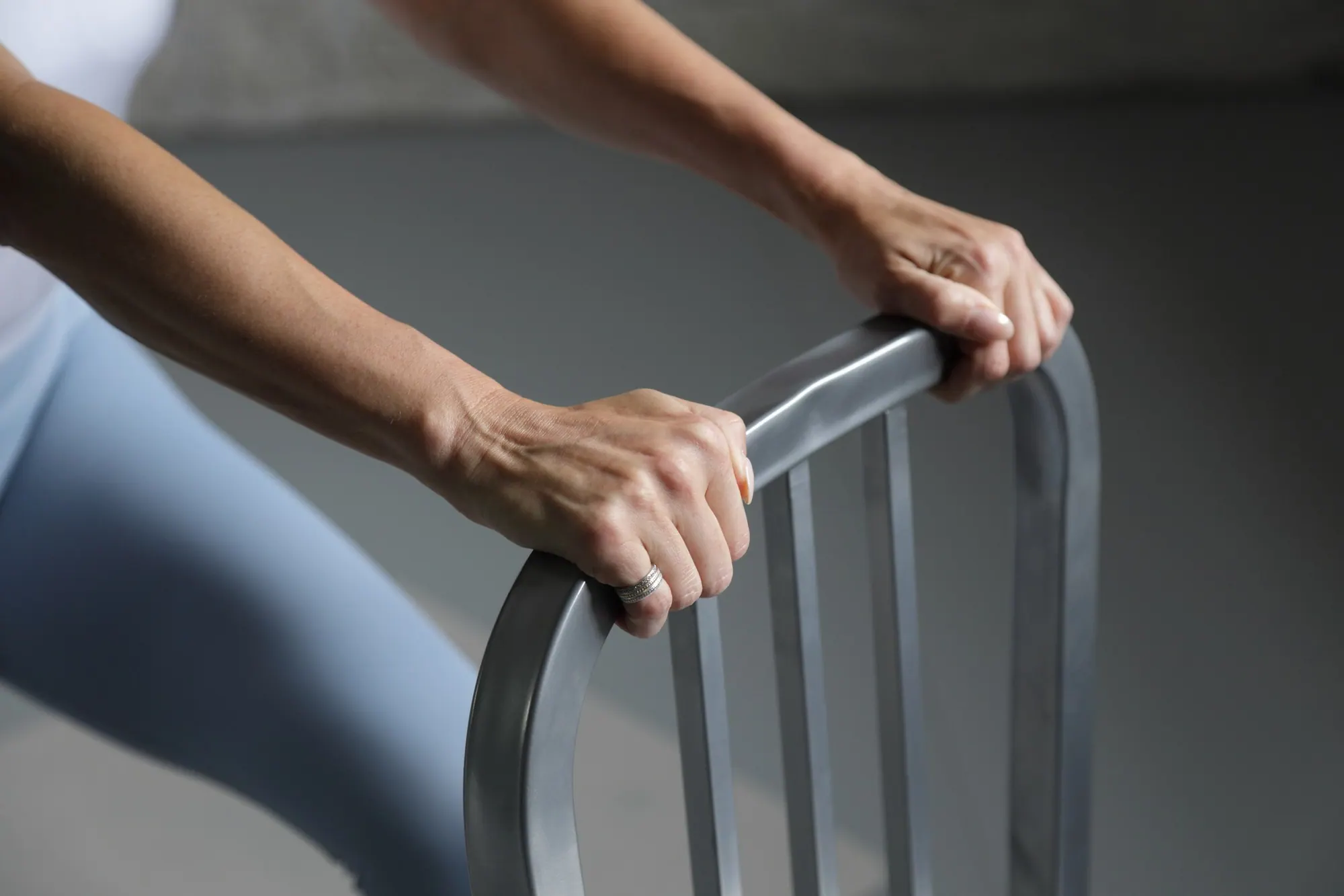 Chrissy’s hand resting on the back of a metal chair, using it as a barre, in a quiet grey studio.